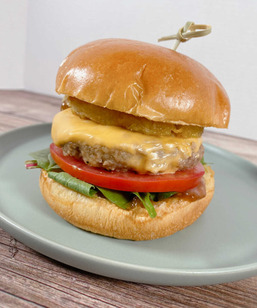 close up image of the burger on a light green plate. From the bottom up: lettuce, tomato, burger, grilled pineapple. Bun held together with a skewer. 