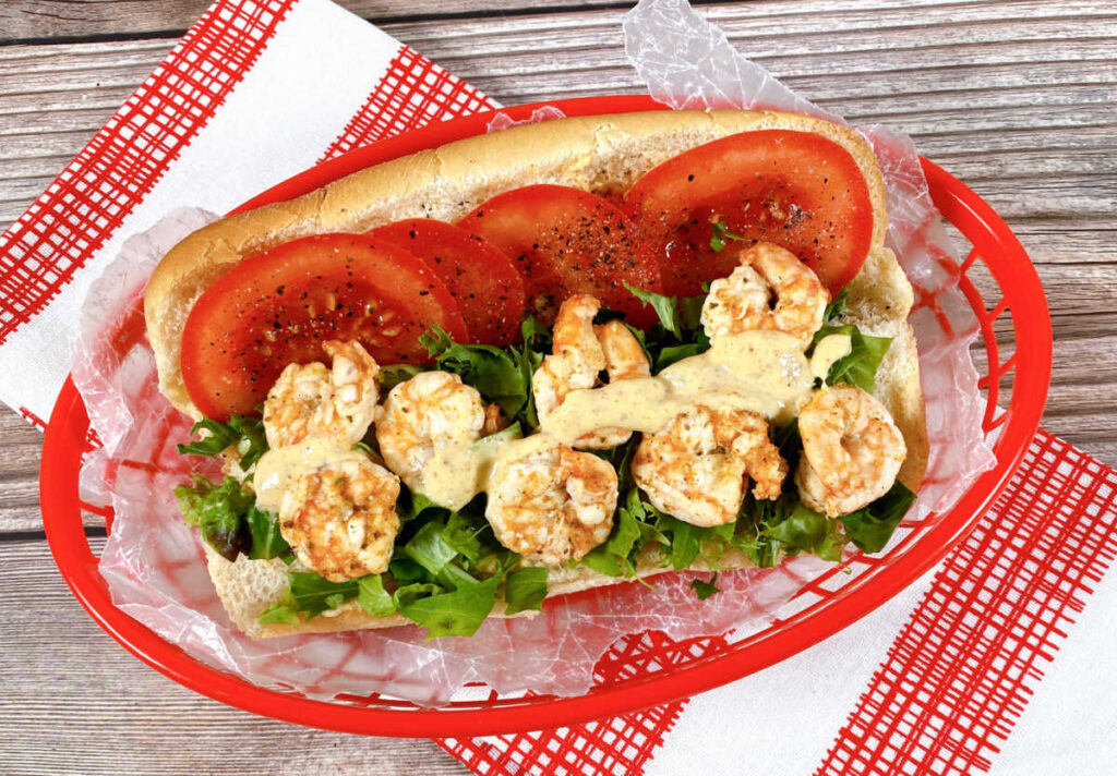 Close up of the sandwich in a red basket on a checkered napkin. The shrimp are drizzled with the remoulade and the tomatoes are sprinkled with pepper. 