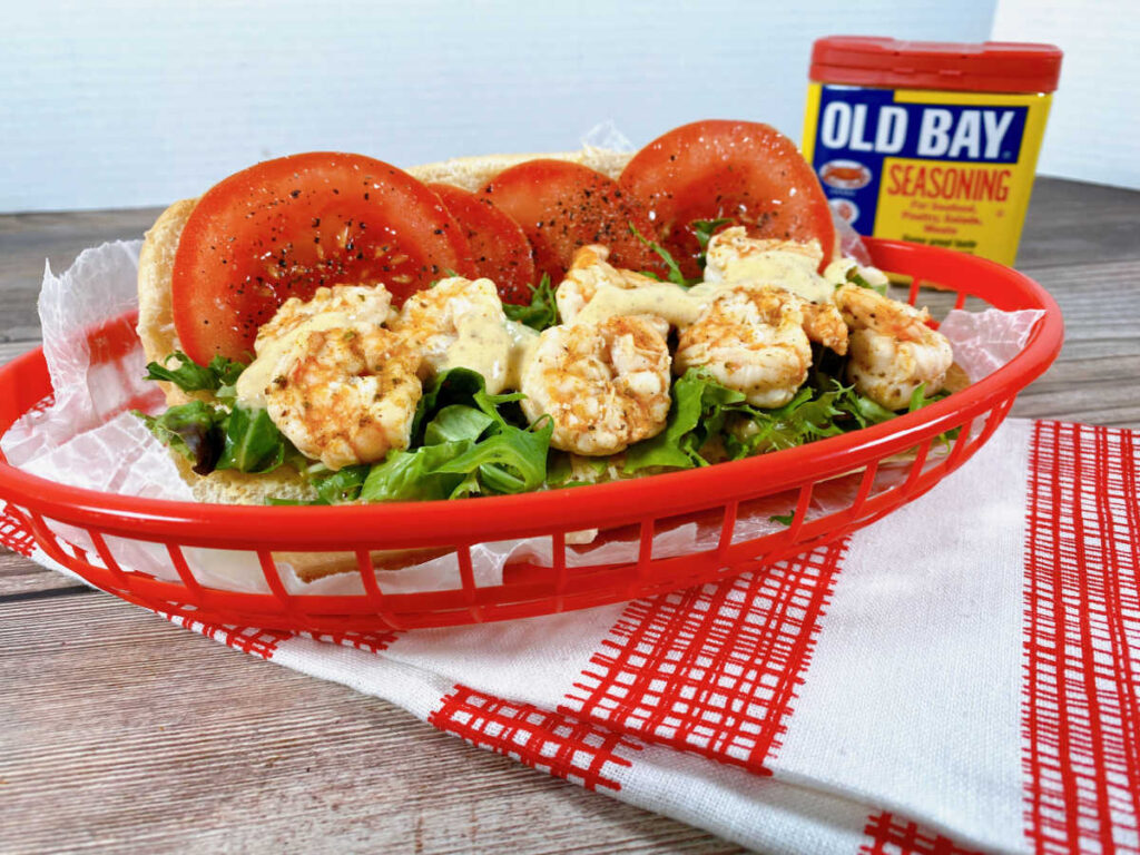 Steamed Shrimp Po Boy sandwich sits in a red basket on a checkered napkin. Shrimp are drizzled with Old Bay Remoulade sauce. 