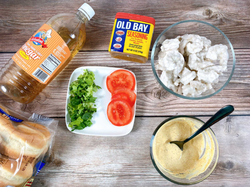 Ingredients for the sandwich sit on a wooden background - shrimp, old bay seasoning, lettuce, tomato slices, rolls, apple cider vinegar and the prepared remoulade.