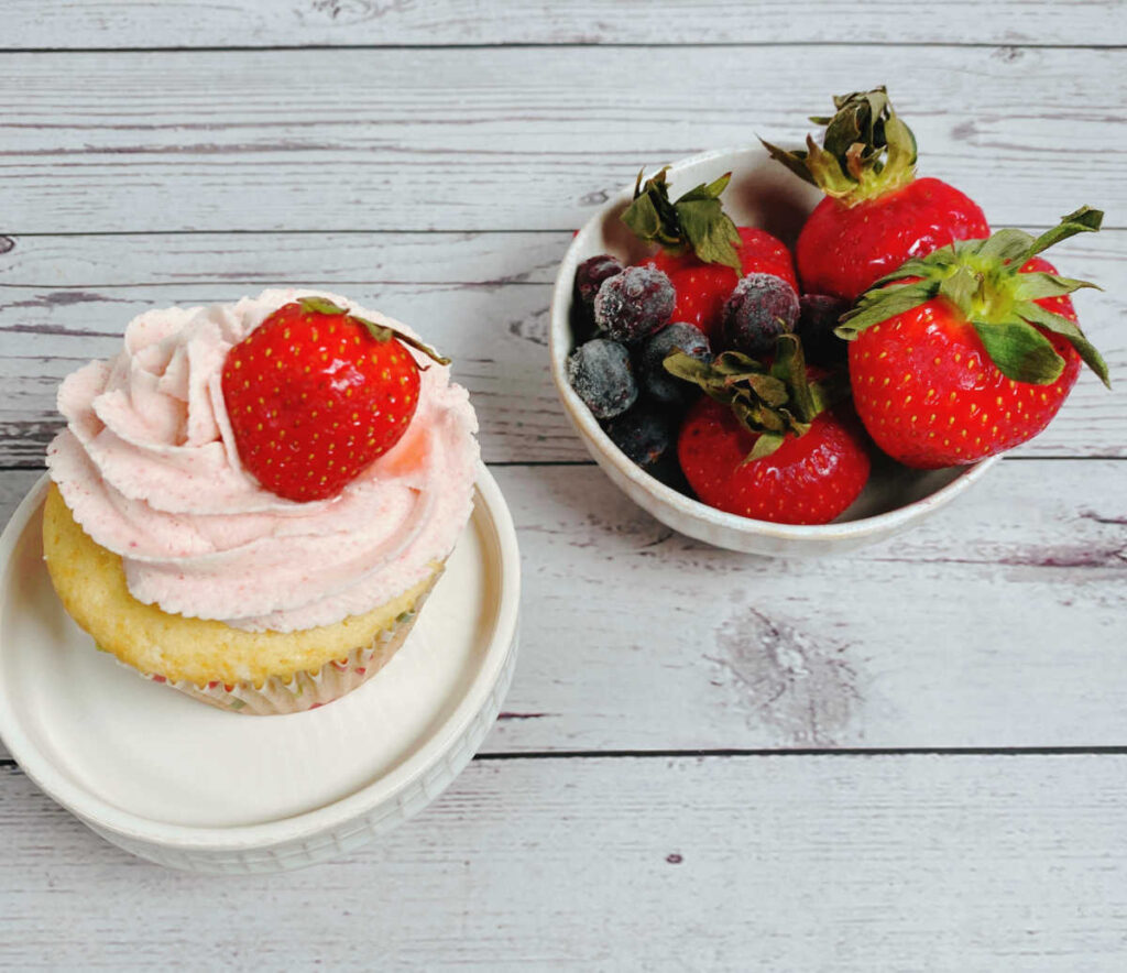 Overhead image of cupcake topped with a strawberry on a cupcake stand. Bowl of strawberries and blueberries is to the right of the cupcake. 