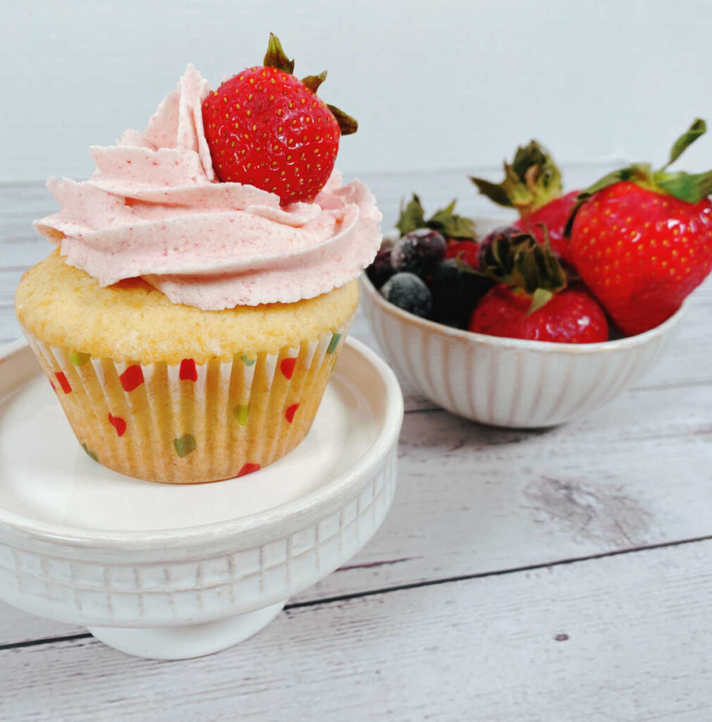 Cupcake topped with a fresh strawberry sits on a white cupcake stand. A bowl of strawberries and blueberries sit in the background. 