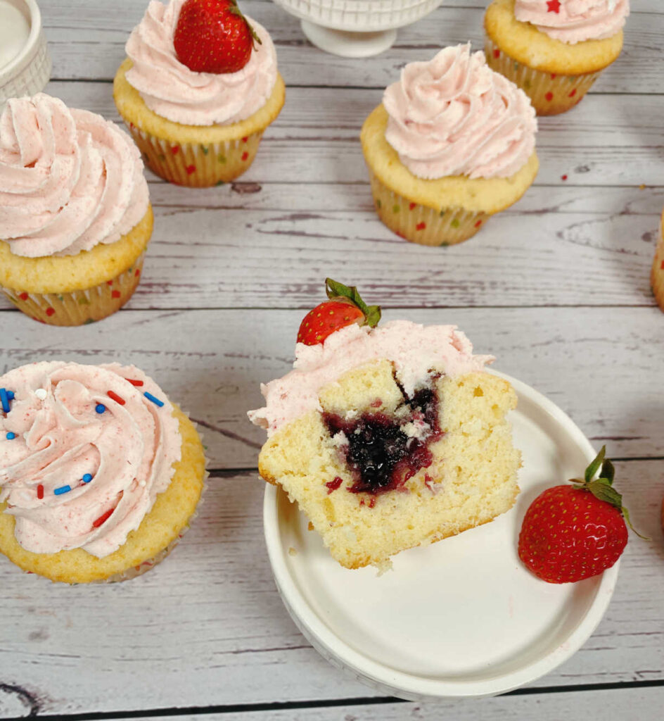 Cupcakes sit on a white wooden background. One cupcake sits on a stand, cut in half lengthwise to show the blueberry jam inside. 