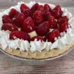 no-bake pie filled with pudding and topped with glaze strawberries and whipped cream sits on a wooden background