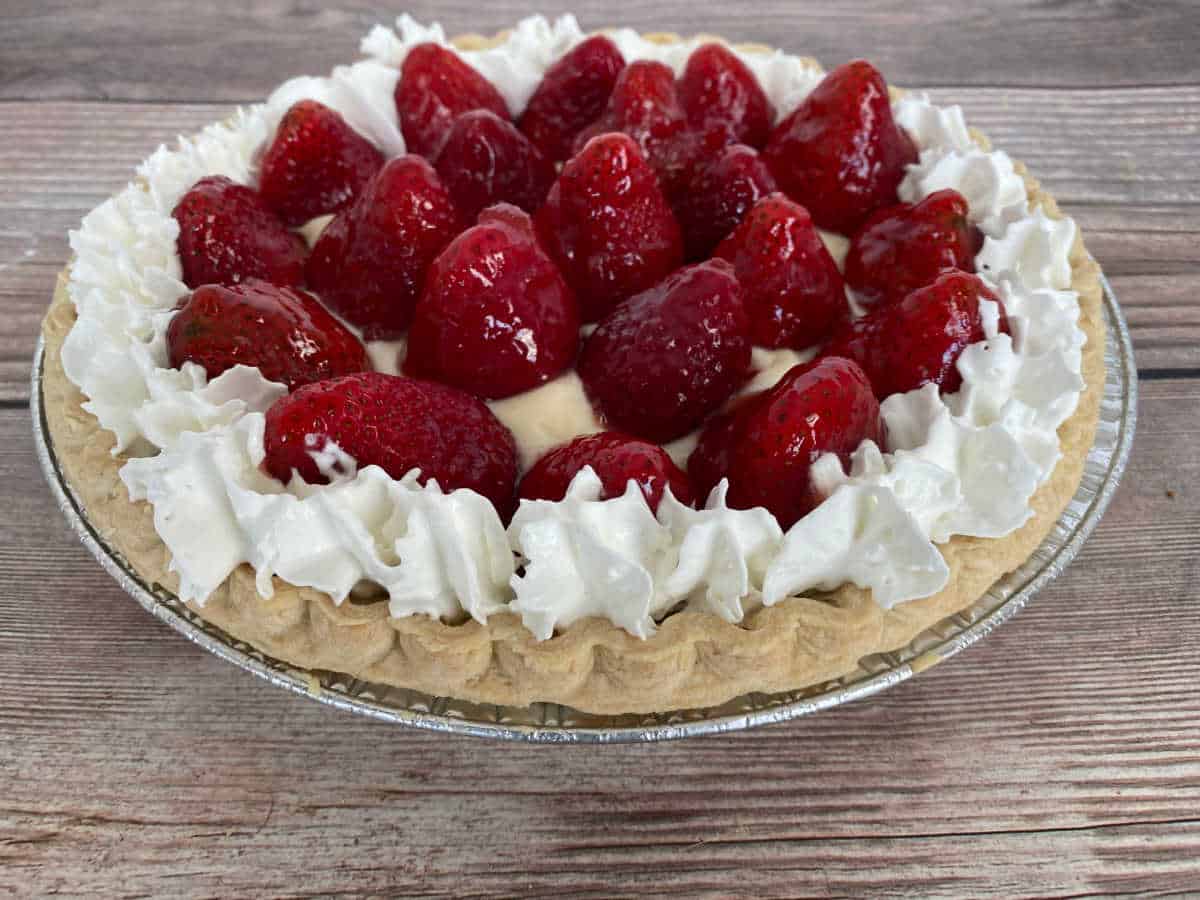 no-bake pie filled with pudding and topped with glaze strawberries and whipped cream sits on a wooden background