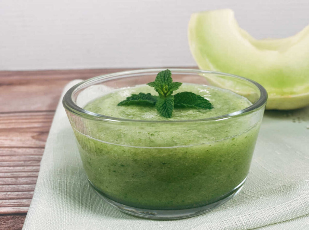 Small glass bowl of the soup sits on a light green napkin with a wedge of honeydew in the background.