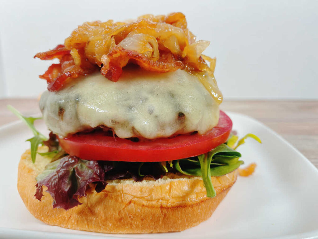 Open faced burger, showcasing the melty cheese, bacon and pile of caramelized onions sits on a white, square plate.