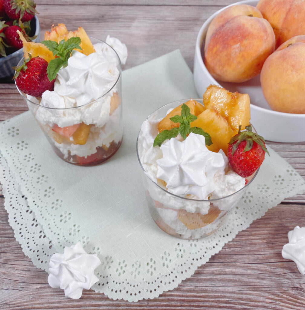 two glasses of eton mess sit on a light green scalloped napkin. A bowl of strawberries and a bowl of peaches sit off to the side, along with meringue cookies.