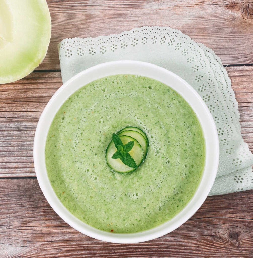 overhead image of soup in a shallow white bowl on a wooden background. Soup is garnished with mint leaves and thinly sliced cucumber.