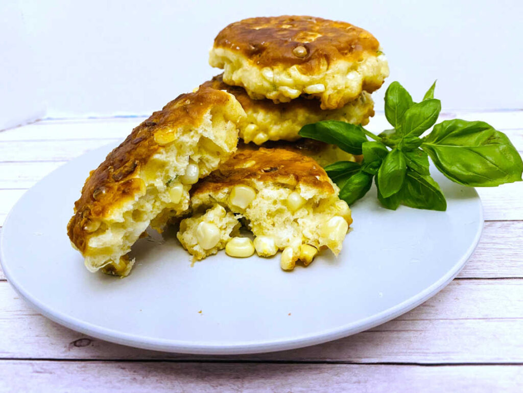 corn fritters sit on a white plate garnished with basil. one fritter is broken open to show all of the corn inside.
