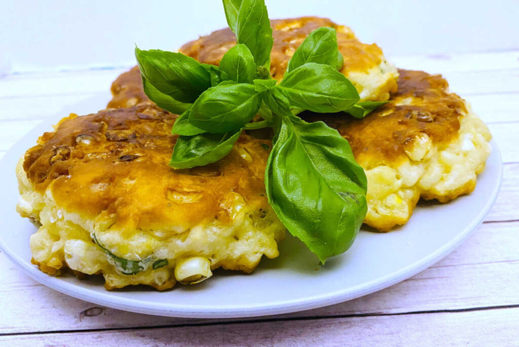 corn fritters are stacked on a white plate and garnished with fresh basil on top. 