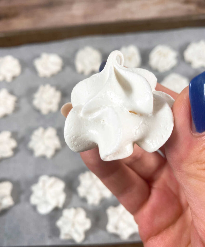 close up of holding a meringue cookie with cookies on the baking sheet below.