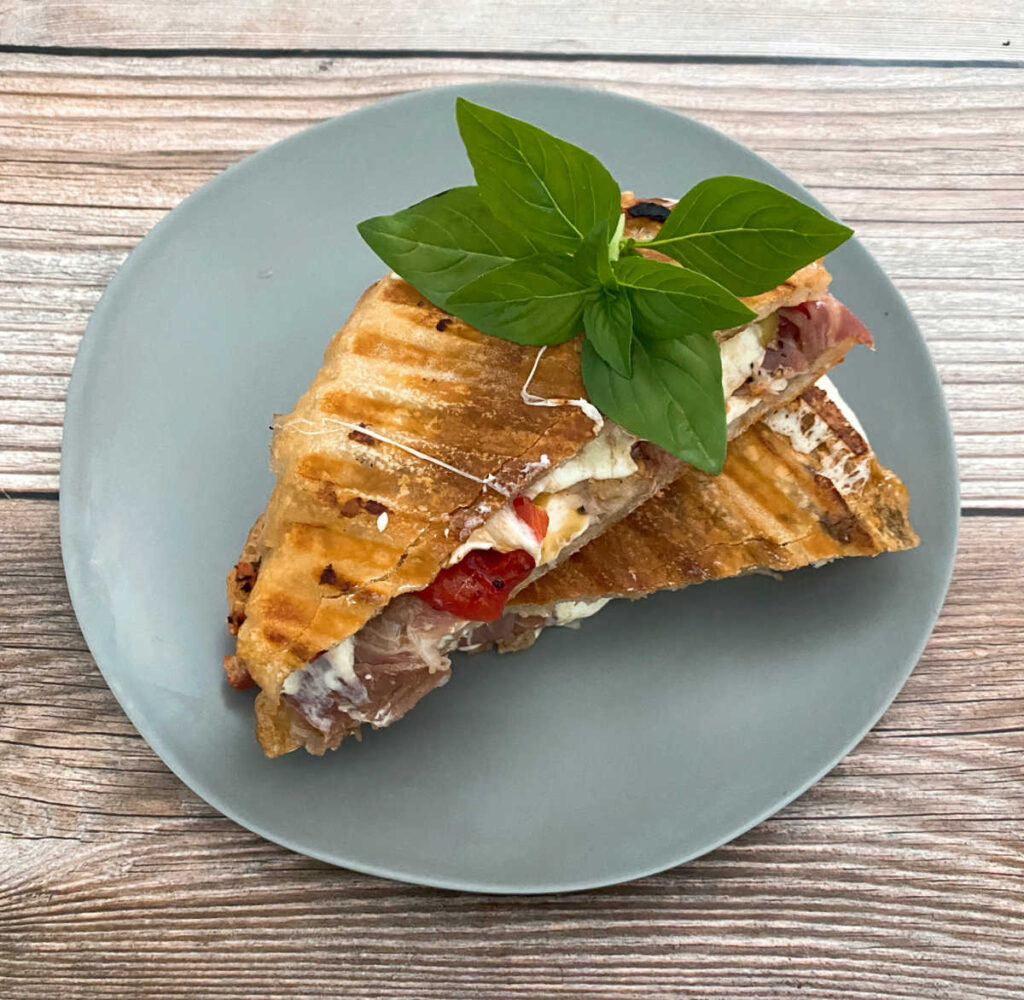 grilled panini, sliced in half and topped with fresh basil, sits on a blue-gray plate on a wooden background