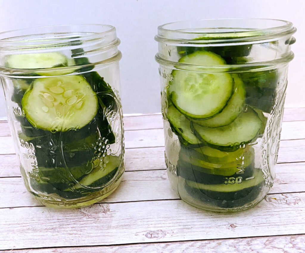 preparation image of the pickle recipe. Cucumber slices are stacked in mason jars waiting for the liquid to cover them. 