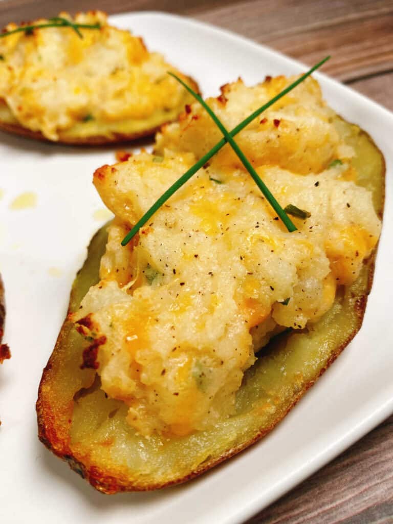 twice baked potatoes, garnished with long strips of fresh chives sit on a white square plate. The filled portion of the potato is cheesy and crispy.