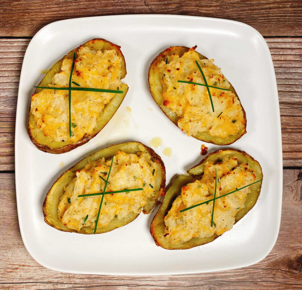 Four potatoes, filled with the creamy, cheesy filling and garnished with long strips of fresh chives sit on a white square plate on a wooden background.