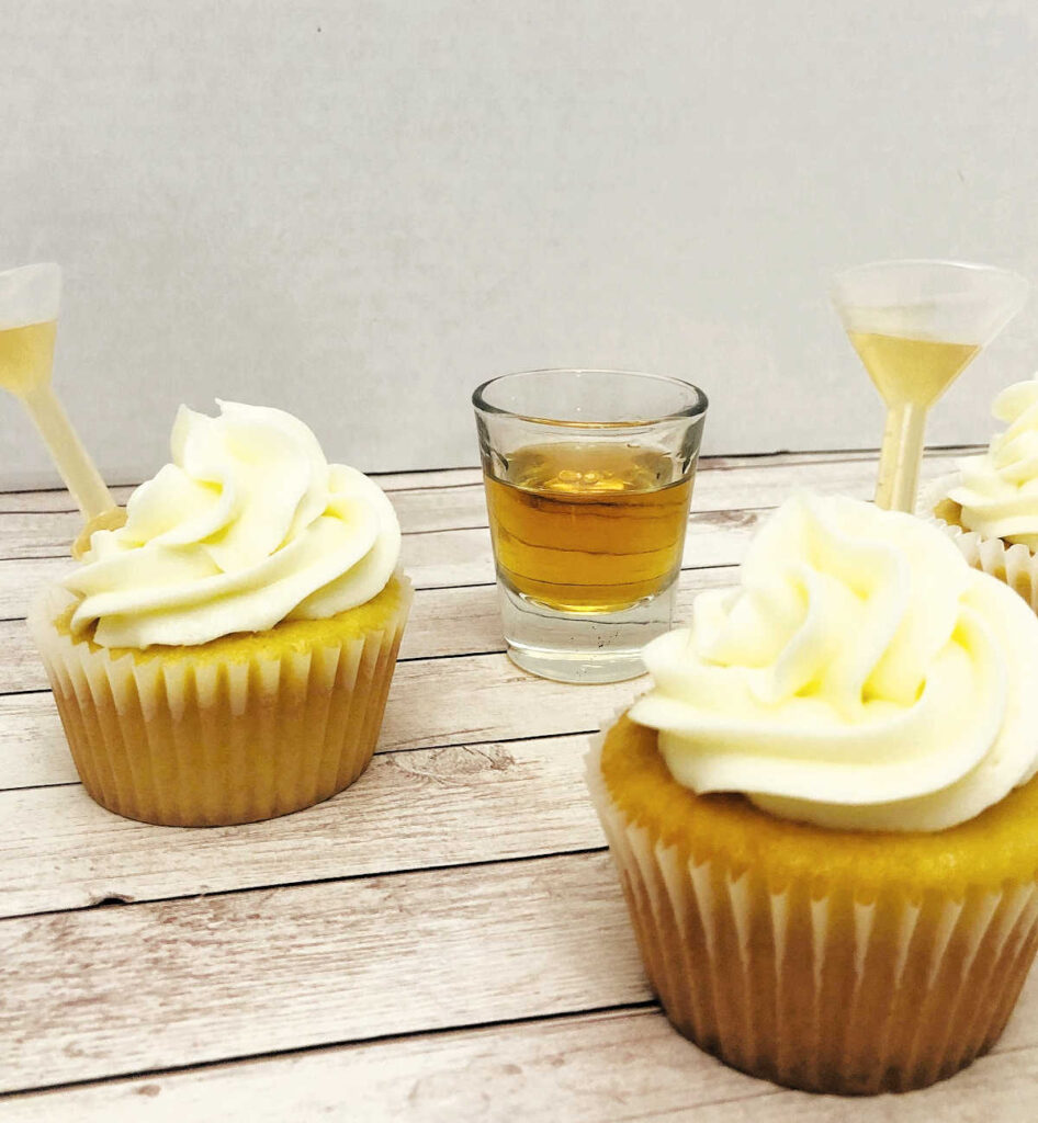 cupcakes filled with bourbon Shot Tops sit on a wooden background. A shot glass of bourbon sits behind the cupcakes. 