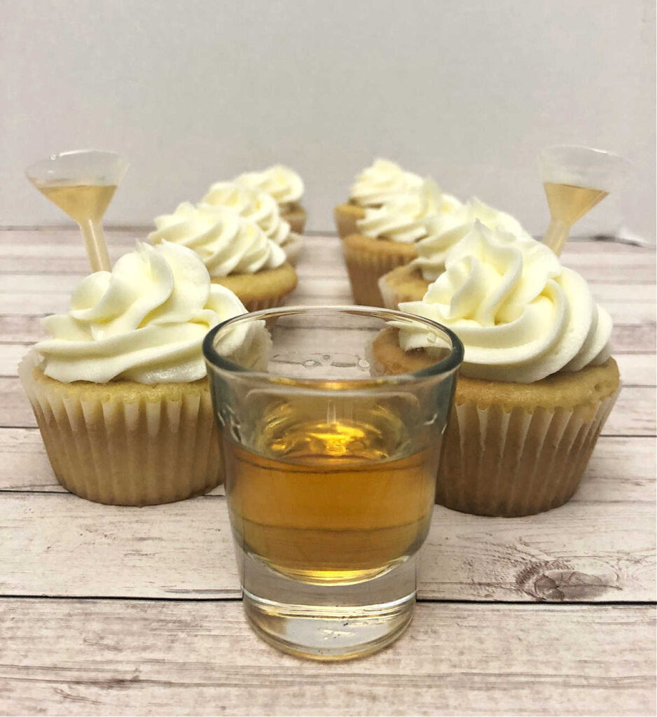 two rows of cupcakes sit on a wooden background with a shot glass of bourbon in front of them. 