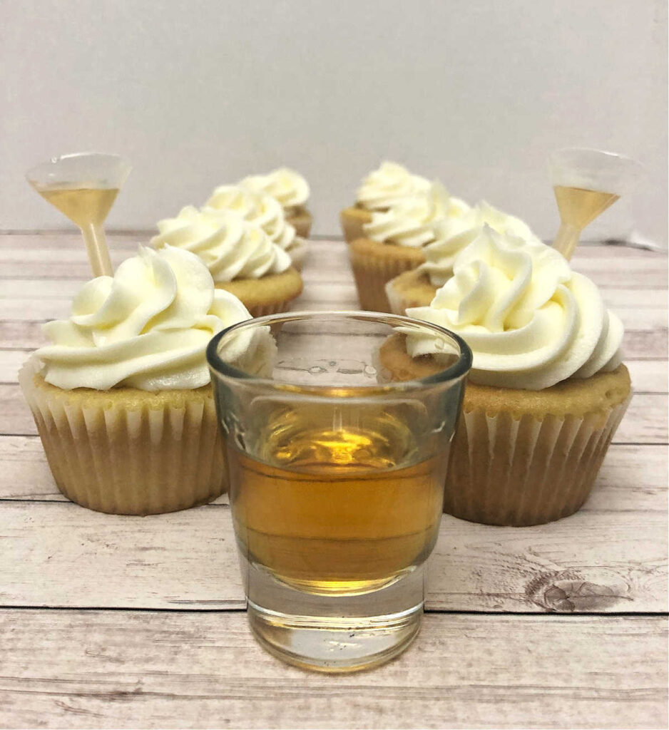 two rows of cupcakes sit on a wooden background with a shot glass of bourbon in front of them.
