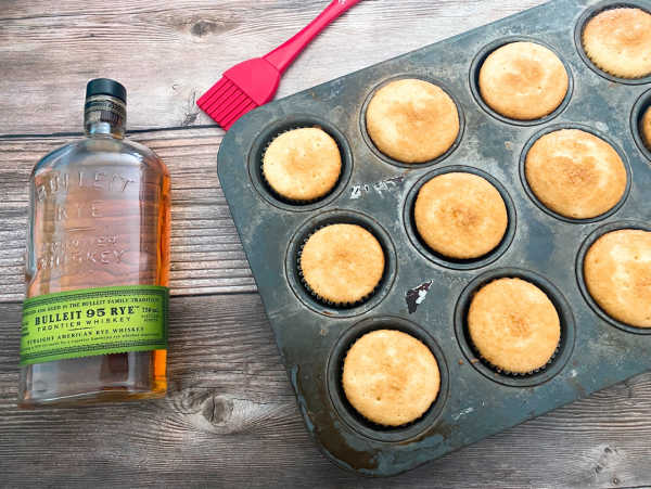 preparation image of recipe - baked cupcakes sit in a cupcake pan with a bottle of bourbon and a pastry brush off to the side, ready to be glazed. 