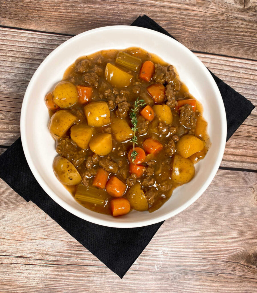 Stew sits in a shallow white bowl on top of a black napkin. Stew is garnished with a sprig of fresh parsley
