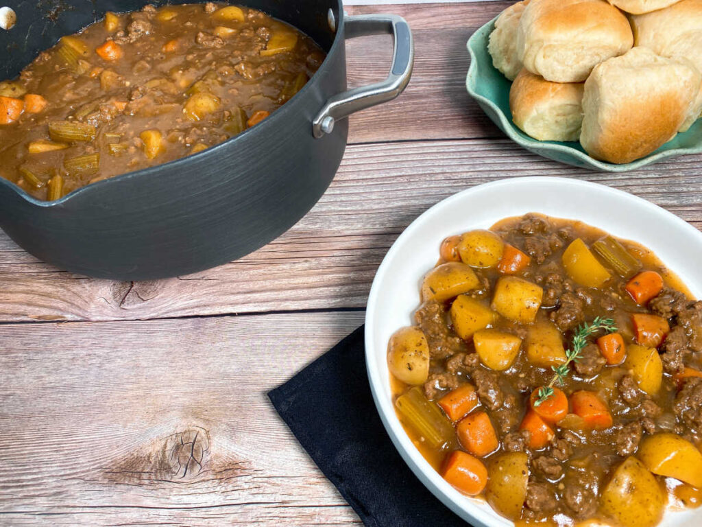 Pot of stew sits to the left, bowl of stew at the bottom and a plate of dinner rolls sit on the right. 