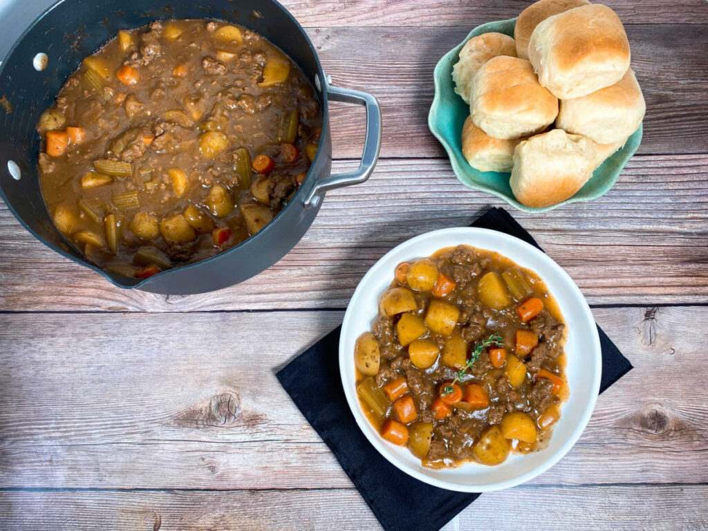 overhead image of stew in pot at the top left. Stew in a shallow white bowl at the bottom and a plate full of dinner rolls on the right. 