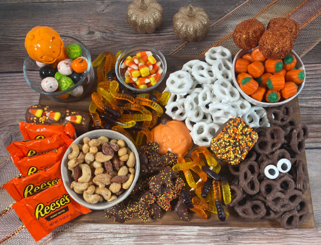 candy, pretzels, gumballs, nuts, marshmallows and other halloween candy sit on a wooden board. Area around the board is decorated with glittery pumpkins.