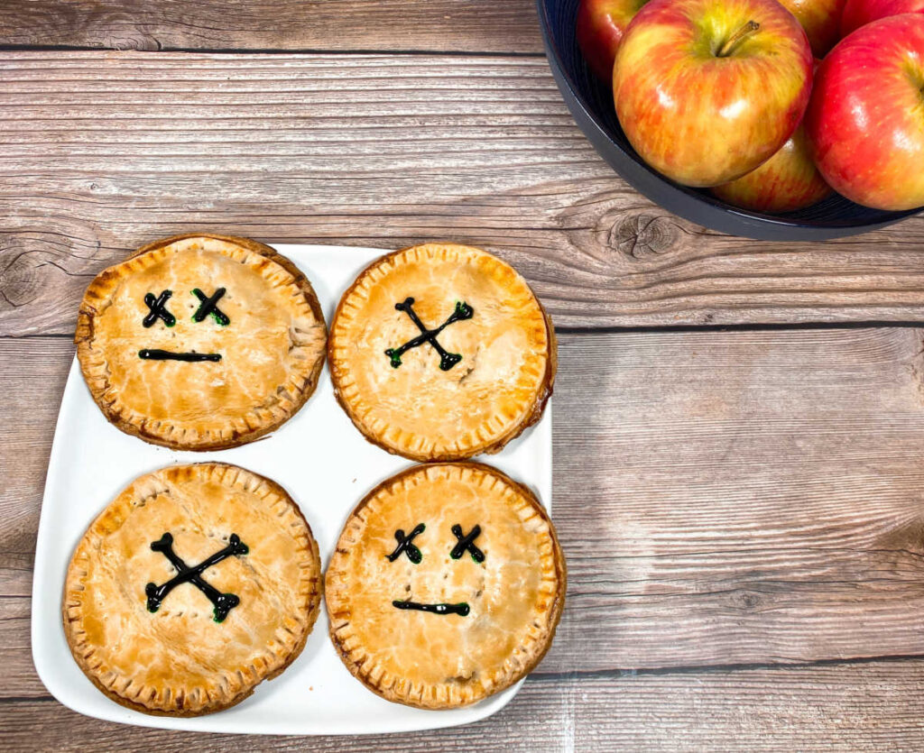 decorated hand pies sit on a white plate on a wooden background with a bowl of apples to the right side of them. 