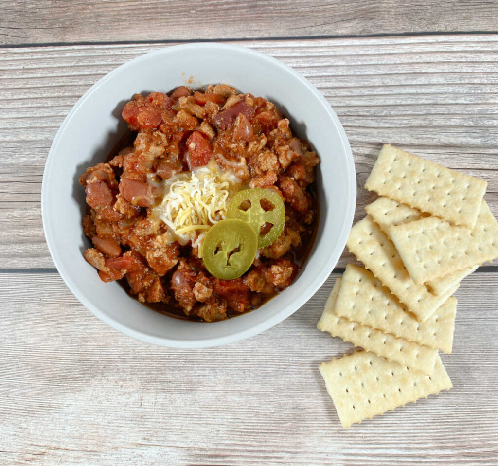 overhead image of turkey chili in a white bowl. Chili is topped with shredded cheese and pickled jalapenos. Crackers sit to the side.