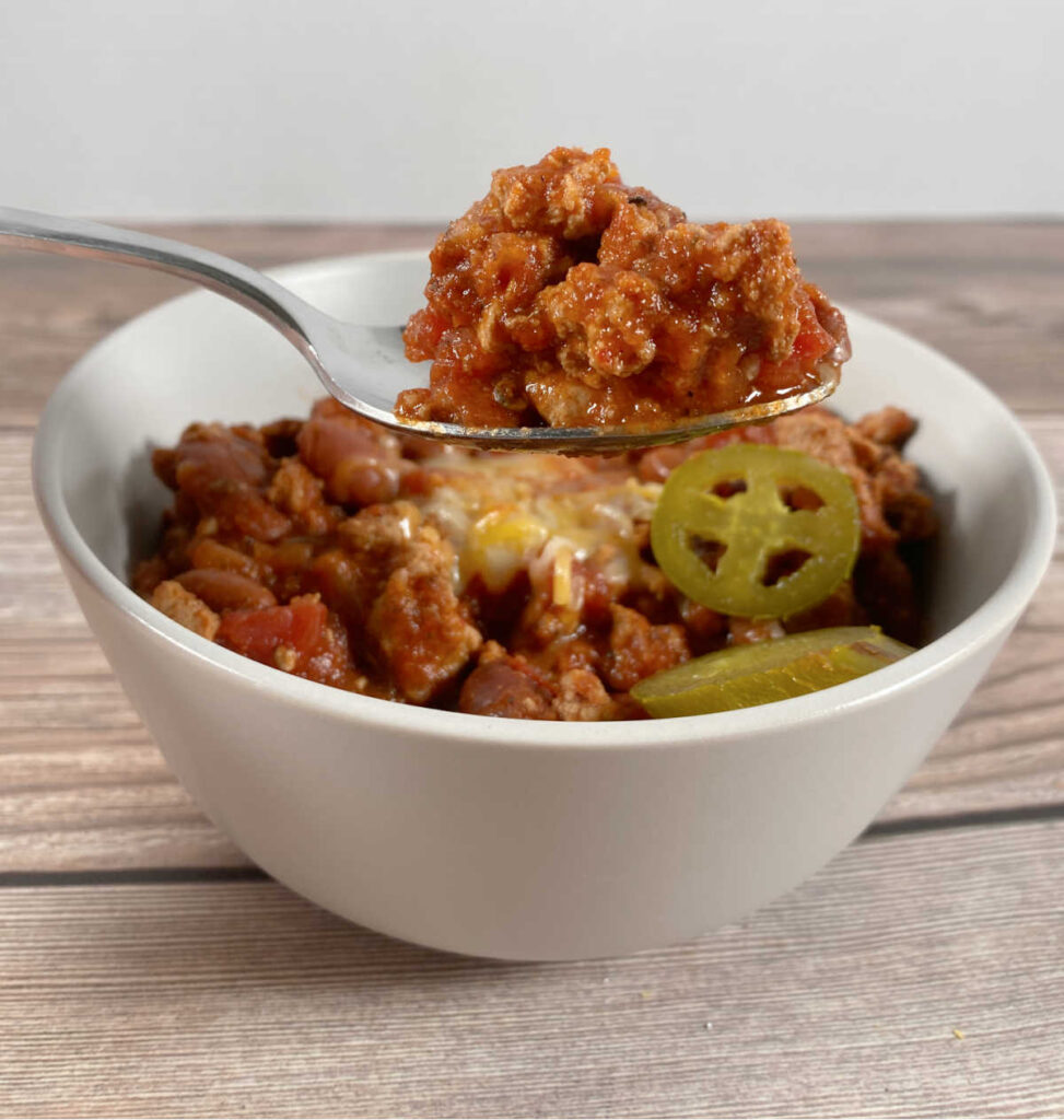 A spoon is lifting up a bite of the turkey chili from the bowl. Melted cheese and pickled jalapenos are visible in the bowl. 