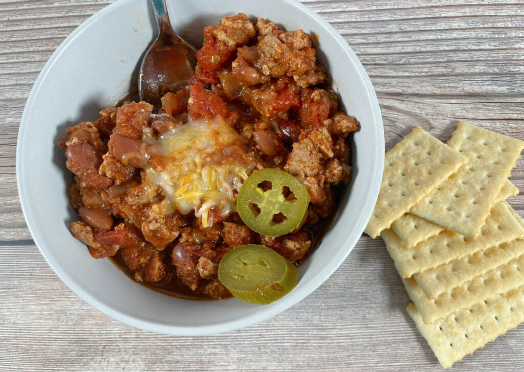 overhead image of the chili in a bowl with melted cheese, pickled jalapenos and crackers on the side. 