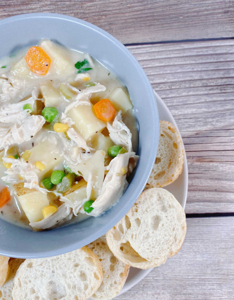 Crock Pot Chicken Pot Pie sits in a pale blue bowl on a wooden background. Soup is surrounded by small bread baguette slices. 