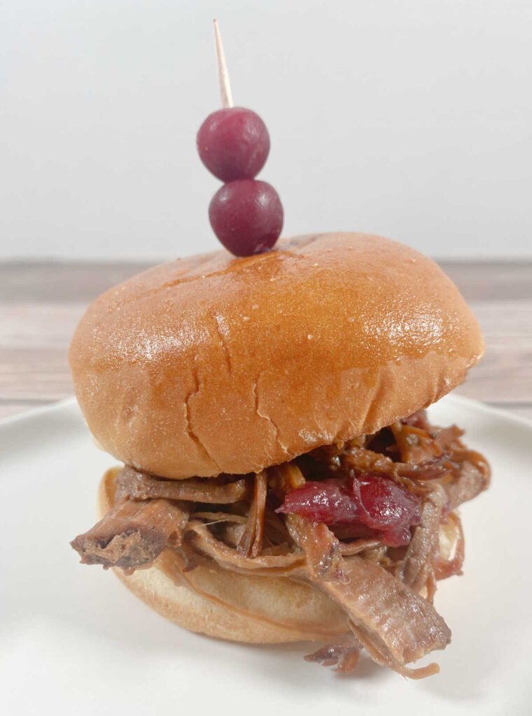 close up of a single slider on a white plate. two cranberries on a toothpick are stuck into the top of the sandwich. 