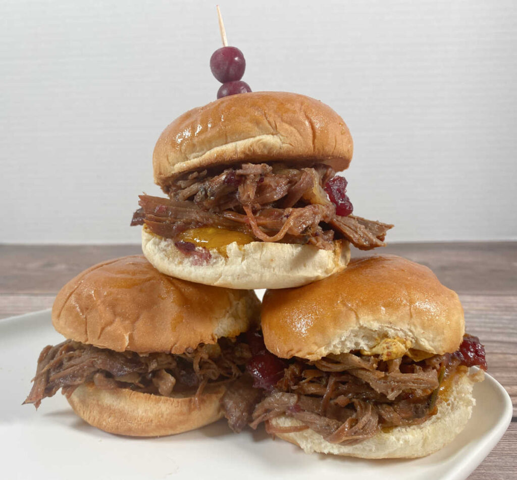 A stack of shredded beef sliders sit on a plate, garnished with fresh cranberries. 