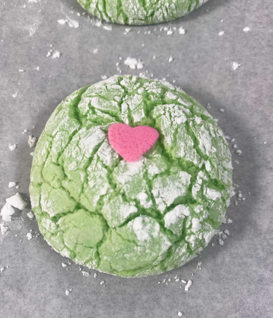 Process shot - baked cookie sits on a baking sheet, heart shaped sprinkle is pressed into the center. 