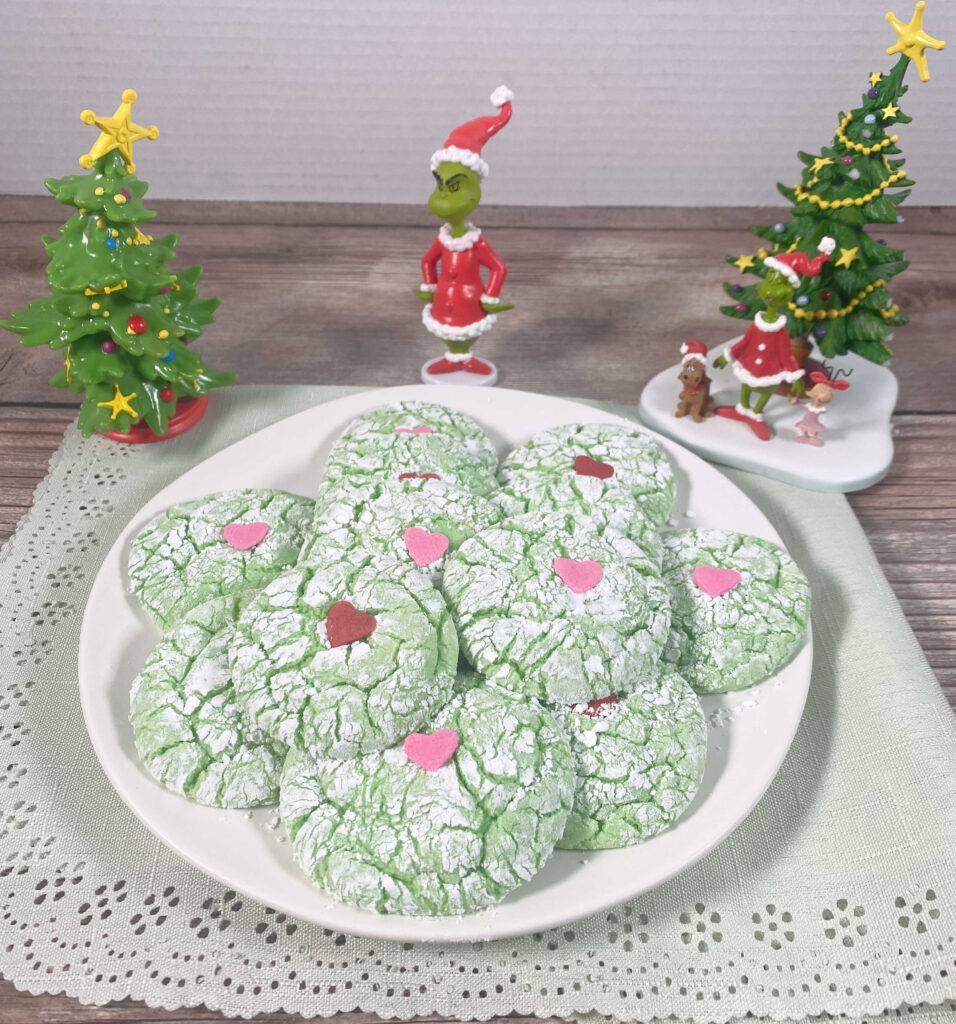 plate of cookies sits on a wooden background with Grinch ornaments surrounding it. 