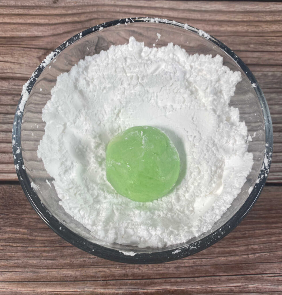 Process shot - green ball of cookie dough sits in a bowl of powdered sugar waiting to be coated. 
