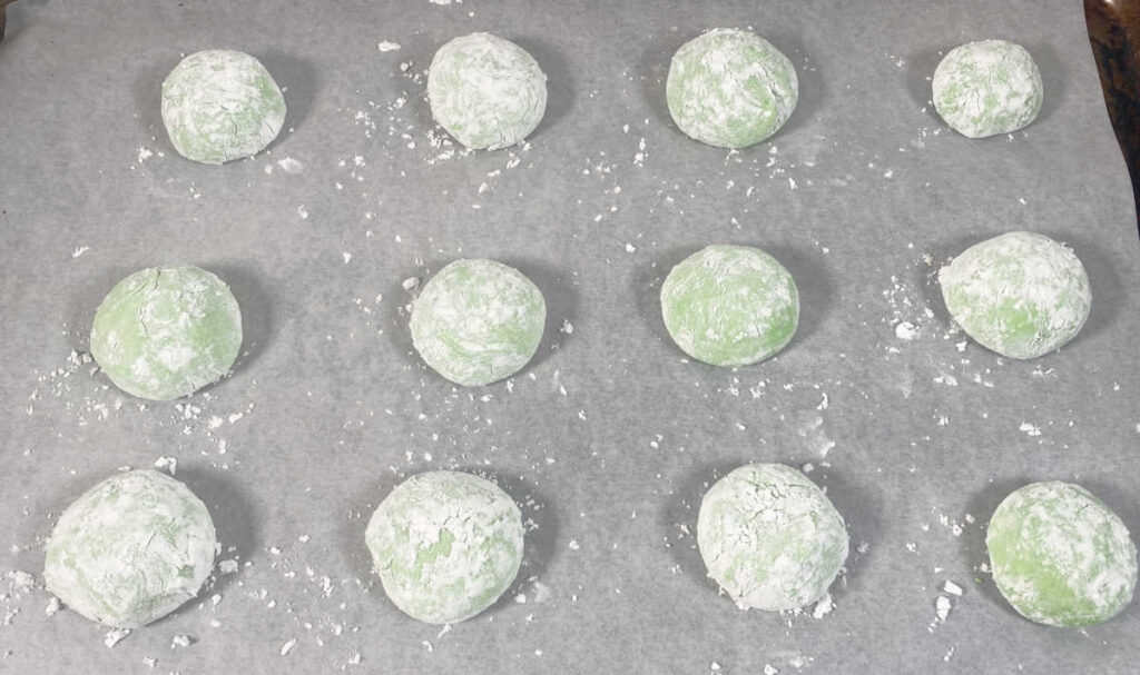 Process shot - powdered sugar coated cookie dough sits on a parchment lined baking sheet ready to be baked. 