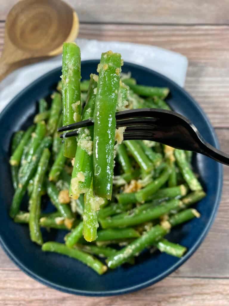 green beans in a dark blue bowl on a wooden background. A fork lifts up some of the beans showing the cheese clinging to them. 