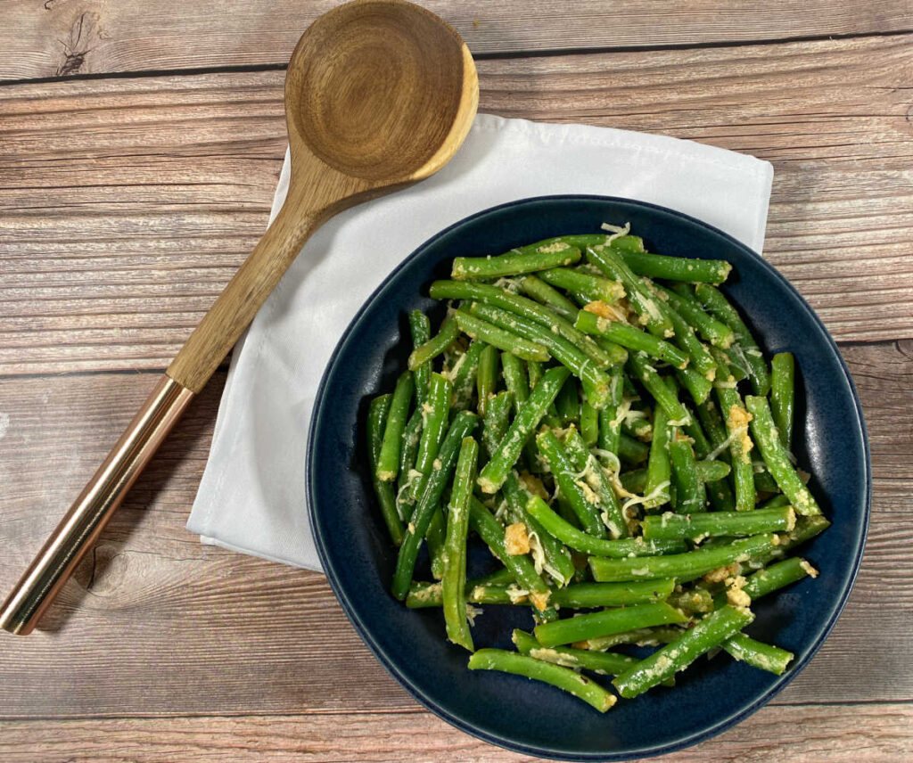 green beans sit in a shallow dark blue bowl, sprinkled with parmesan cheese. a wooden spoon sits to their left on a light green napkin.