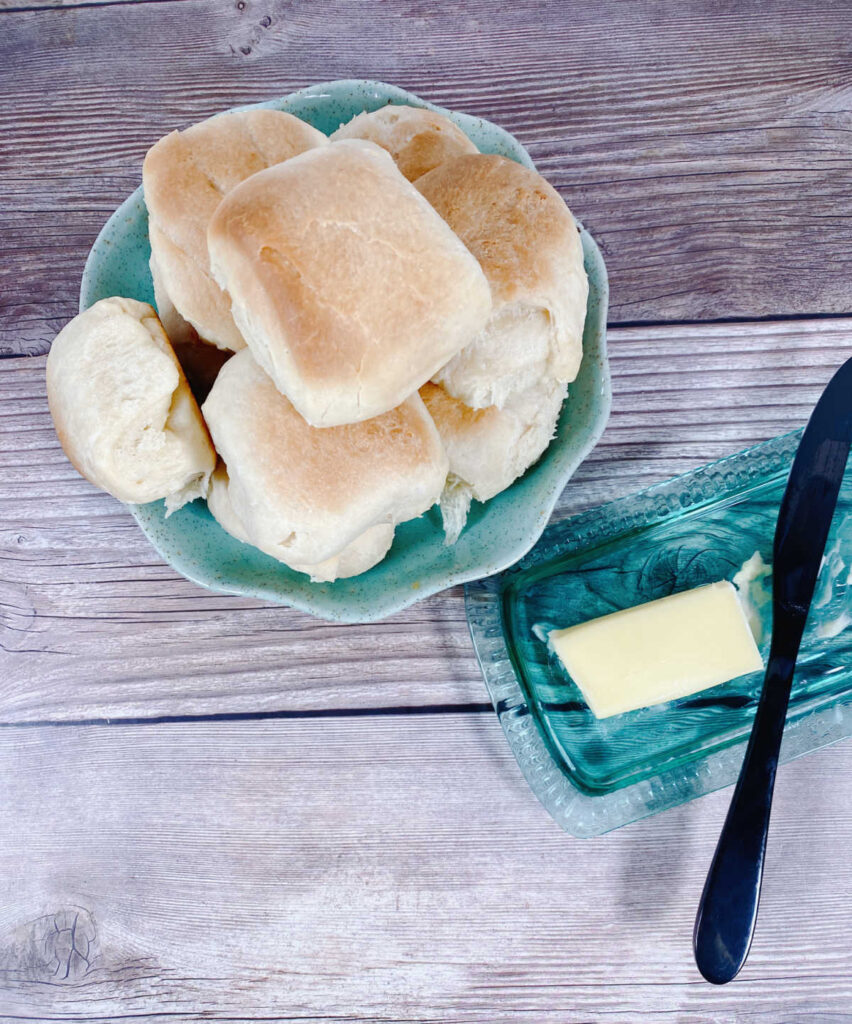 a pile of dinner rolls sit in a round turquoise dish on a wooden background with a butter dish off to the right side. 