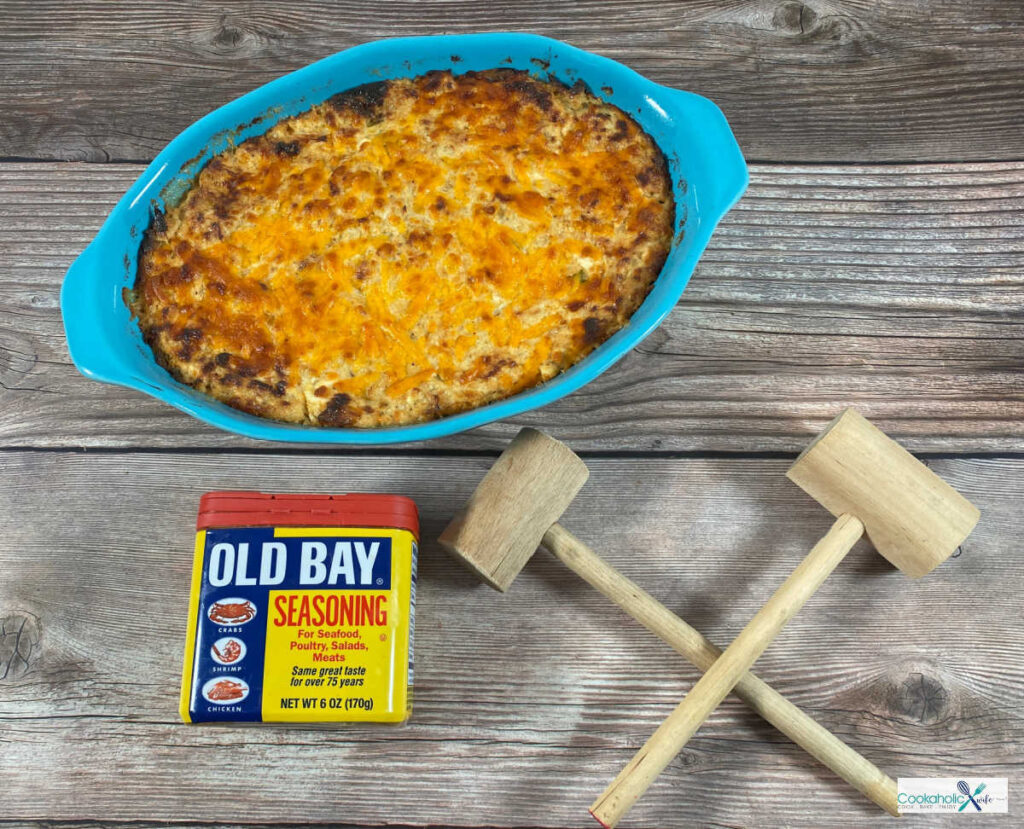 serving dish of crab dip sits on a wooden background. A can of Old bay some some crab mallets sit in the foreground.