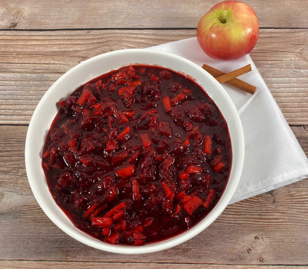 cranberry sauce sits in a shallow white bowl on a wooden background. cinnamon sticks and an apple sit off to the side.
