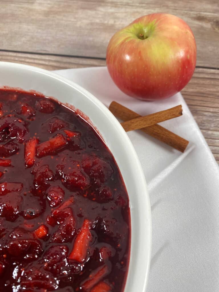 close up image of cranberry sauce in a white bowl with an apple and some cinnamon sticks as decor. 