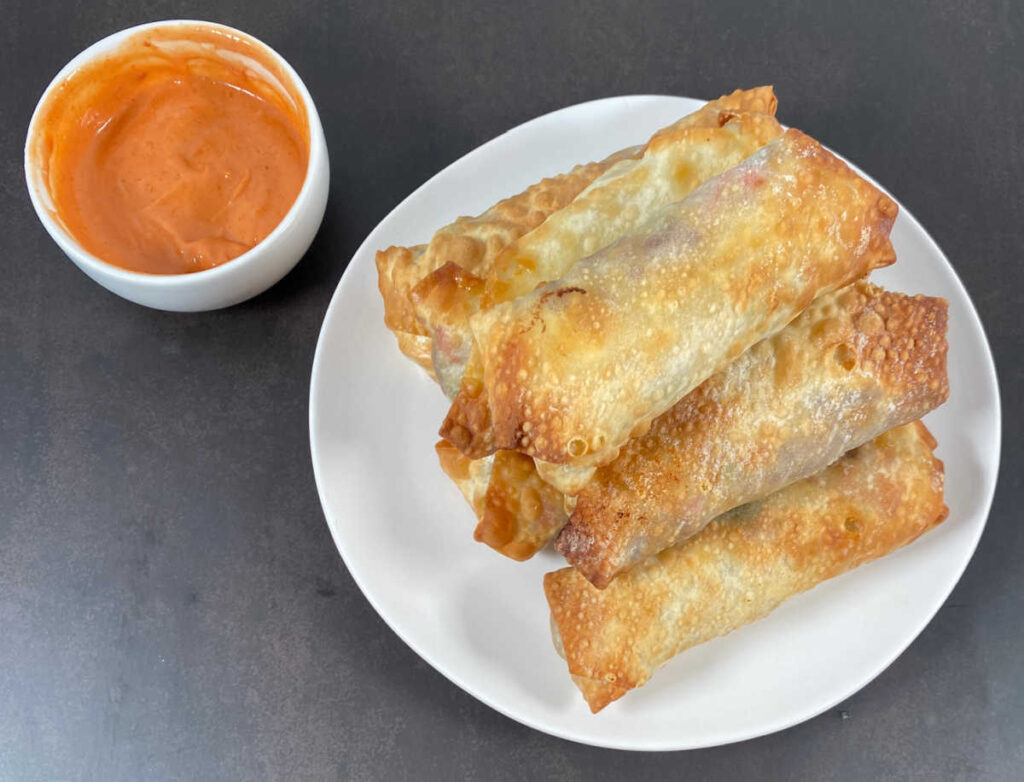 Pyramid of air fried egg rolls sits on a white plate with a bowl of dipping sauce in the top left of the image. 