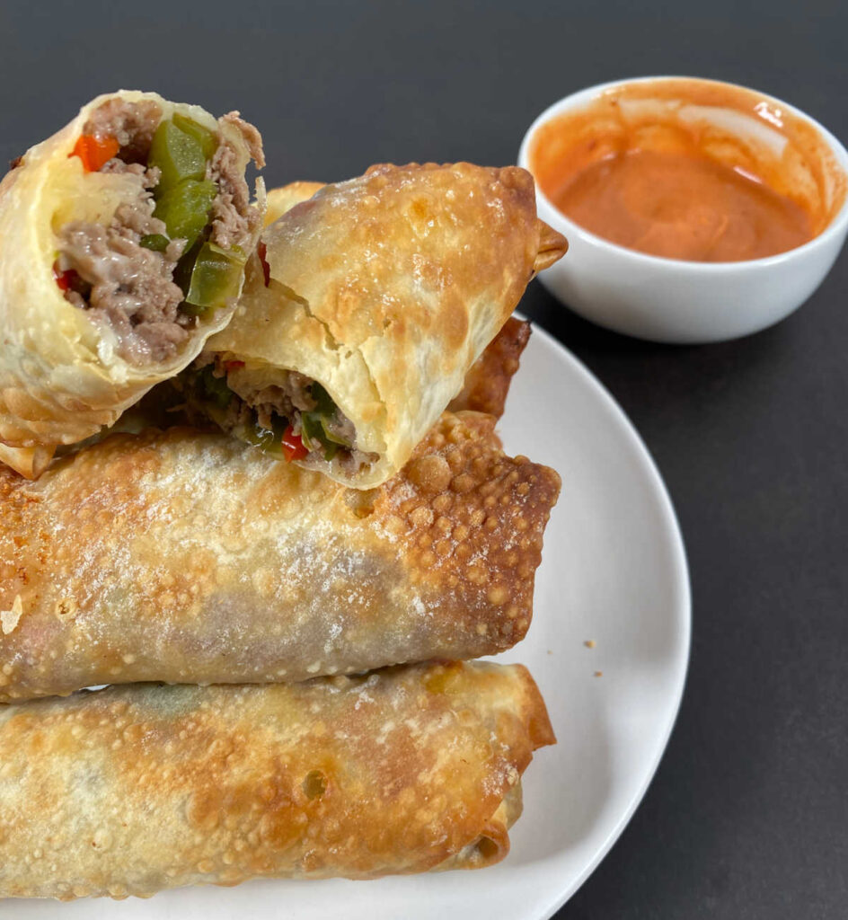 Pyramid of egg rolls with the top one cut in half to show the filling sits on a white plate. A  bowl of dipping sauce sits to the top right. 