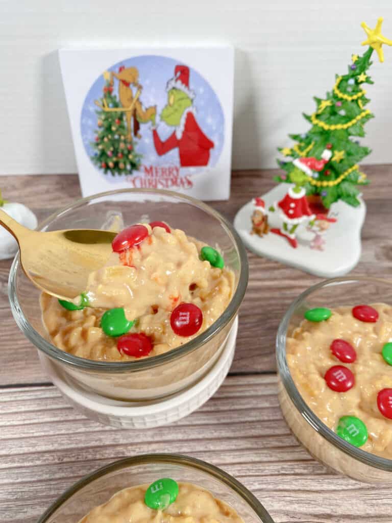 Bowls of pudding sit in glass jars on a wooden background, surrounded with Grinch decor. A spoon is lifting a serving of the pudding out of the bowl. 