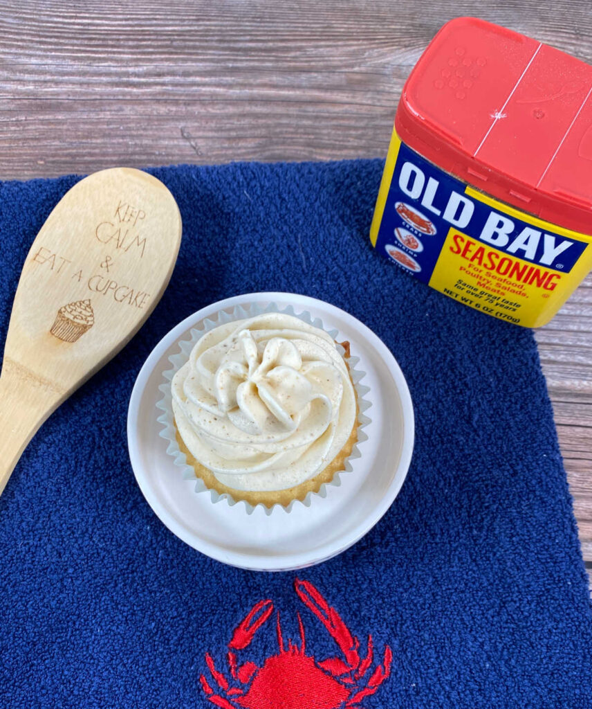 Overhead image of cupcake on a stand, sitting on a blue kitchen towel. A decorative spoon sits to the left, the can of old bay to the right. 