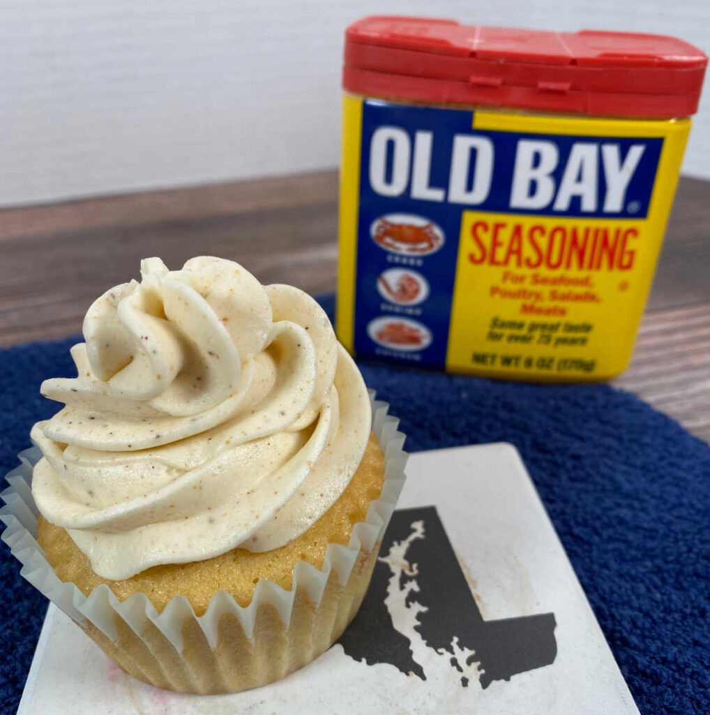 Close up image of cupcake sitting on a white coaster with the state of Maryland outline. Can of old bay sits in the background. 